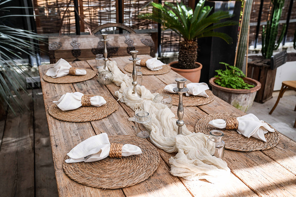 Dekorierter Holztisch mit 6 Tischsets aus Wasserhyazinthe, Kerzenständer, Tischläufer, Vasen und Servietten mit Serviettenring aus Rattan. Pflanzen im Hintergrund.