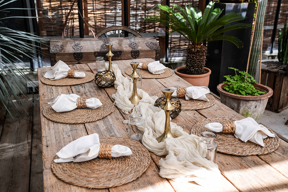 Dekorierter Holztisch mit 6 Tischsets aus Wasserhyazinthe, Tischläufer, Kerzenständer, Vasen und Servietten mit Serviettenring aus Rattan. Pflanzen im Hintergrund.