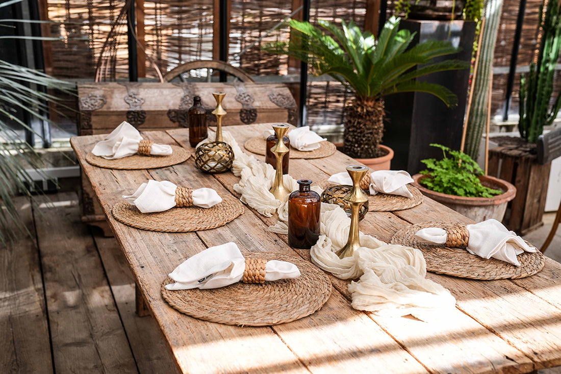 Dekorierter Holztisch mit 6 Tischsets aus Wasserhyazinthe, Tischläufer, Kerzenständer, Vasen und Servietten mit Serviettenring aus Rattan. Pflanzen im Hintergrund.