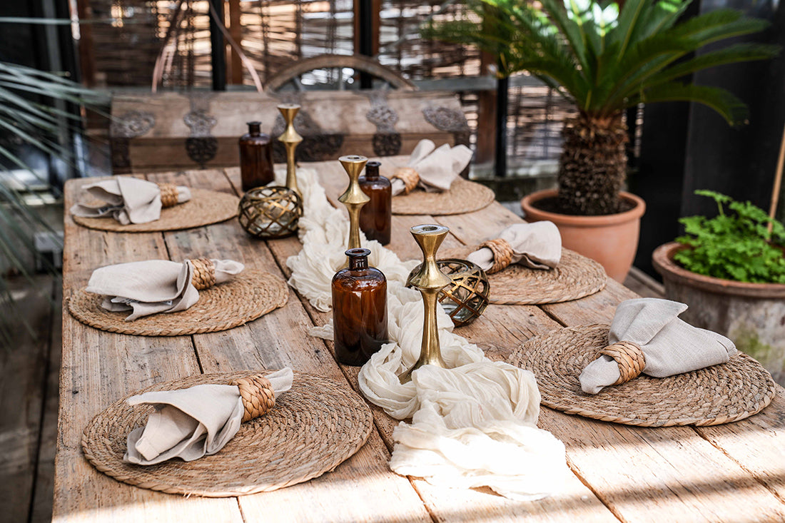 Dekorierter Holztisch mit 6 Tischsets aus Wasserhyazinthe, Tischläufer, Kerzenständer, Vasen und Servietten mit Serviettenring aus Rattan. Pflanzen im Hintergrund.