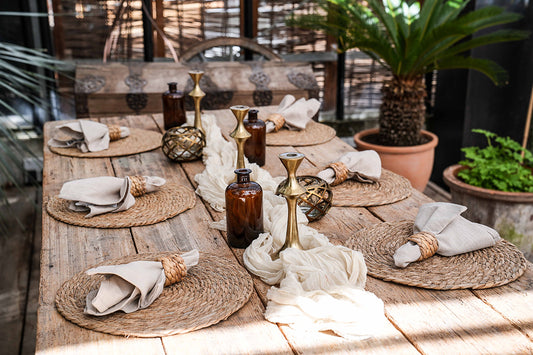 Dekorierter Holztisch mit 6 Tischsets aus Wasserhyazinthe, Tischläufer, Kerzenständer, Vasen und Servietten mit Serviettenring aus Rattan. Pflanzen im Hintergrund.