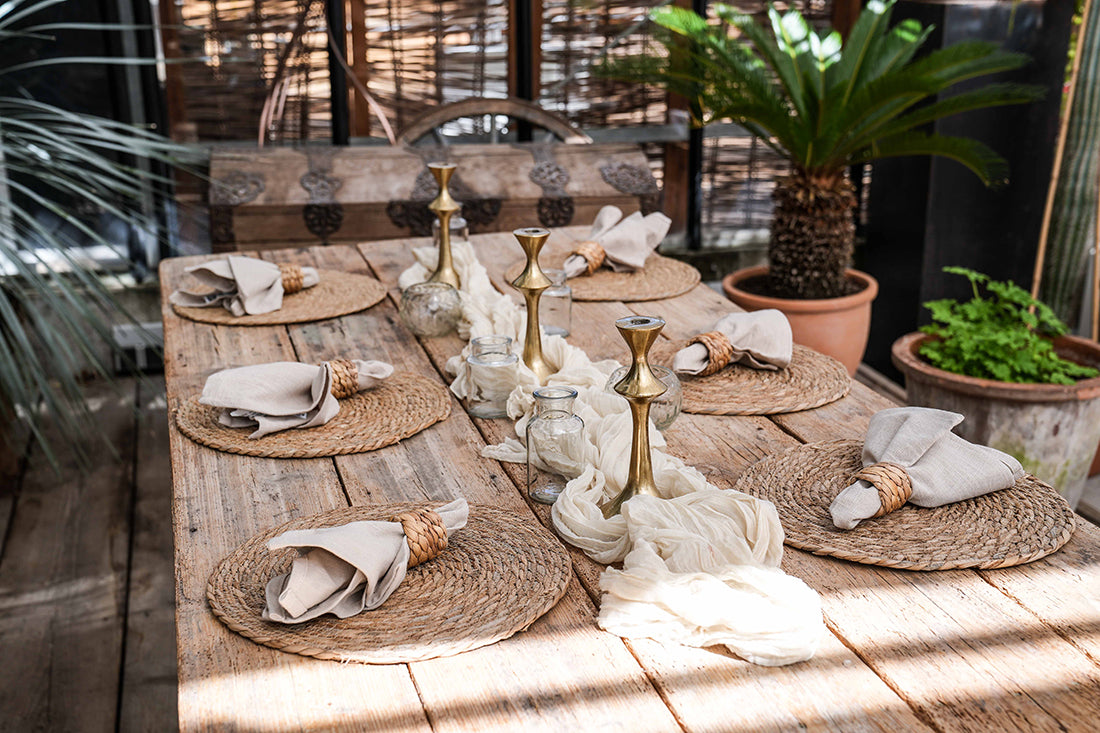 Dekorierter Holztisch mit 6 Tischsets aus Wasserhyazinthe, Tischläufer, Kerzenständer, Vasen und Servietten mit Serviettenring aus Rattan. Pflanzen im Hintergrund.