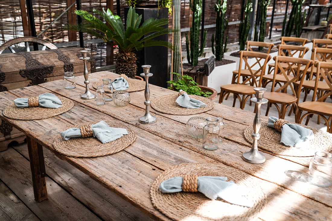 Dekorierter Holztisch mit 6 Tischsets aus Wasserhyazinthe, Tischläufer, Kerzenständer, Vasen und Servietten mit Serviettenring aus Rattan. Pflanzen im Hintergrund.