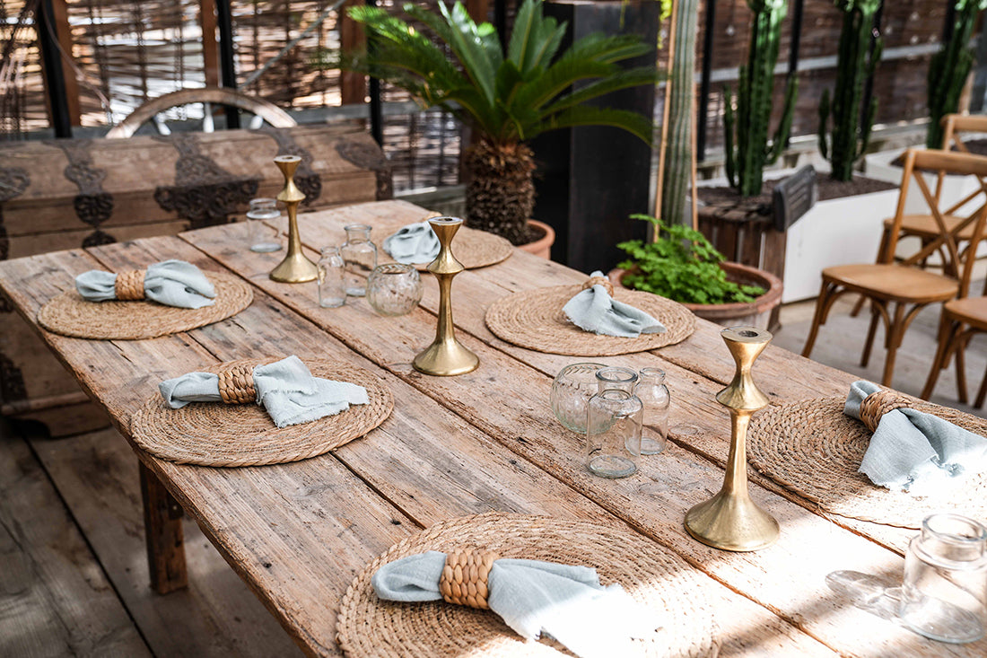 Dekorierter Holztisch mit 6 Tischsets aus Wasserhyazinthe, Tischläufer, Kerzenständer, Vasen und Servietten mit Serviettenring aus Rattan. Pflanzen im Hintergrund.