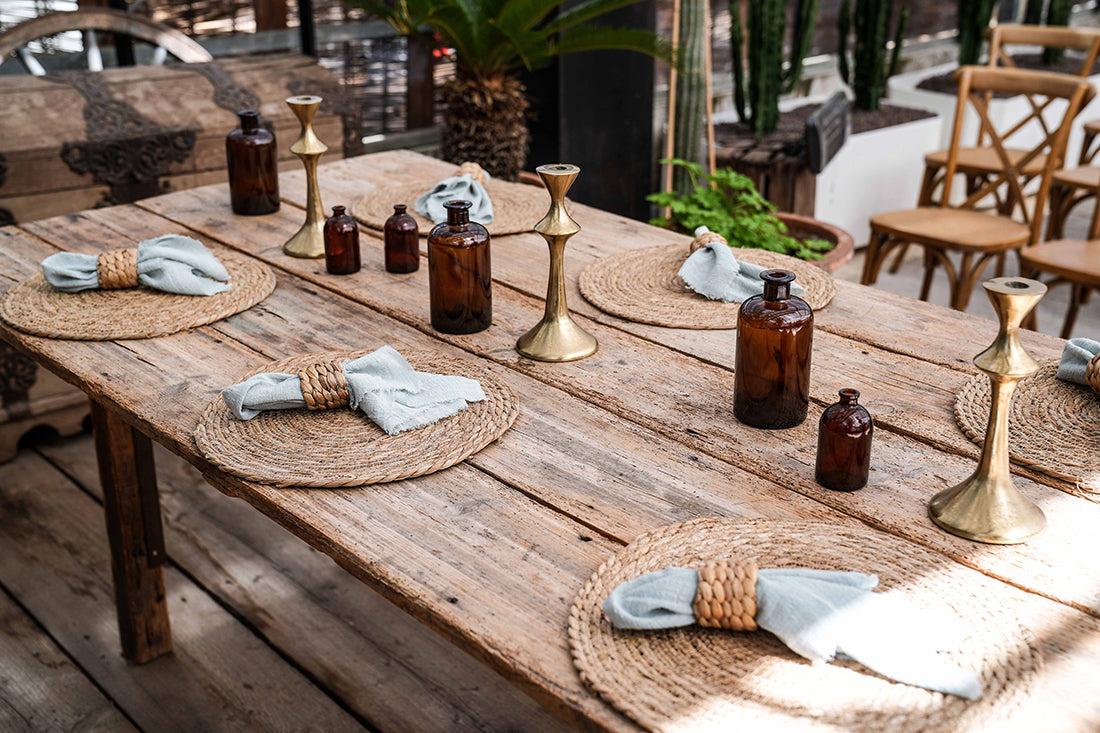 Dekorierter Holztisch mit 6 Tischsets aus Wasserhyazinthe, Tischläufer, Kerzenständer, Vasen und Servietten mit Serviettenring aus Rattan. Pflanzen im Hintergrund.